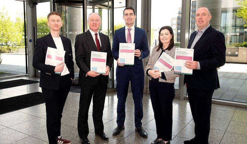 Five people stand indoors, smiling and holding reports. They are dressed in business attire. Large windows and a bright day are visible outside.