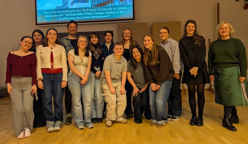 A diverse group of people stands smiling on a wooden floor in front of a screen displaying a welcome message. The atmosphere is warm and cheerful.