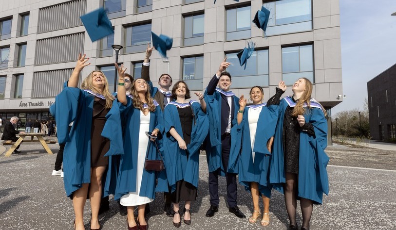 A group of graduates in blue gowns joyfully toss their caps into the air outside a modern building, celebrating their achievement together.