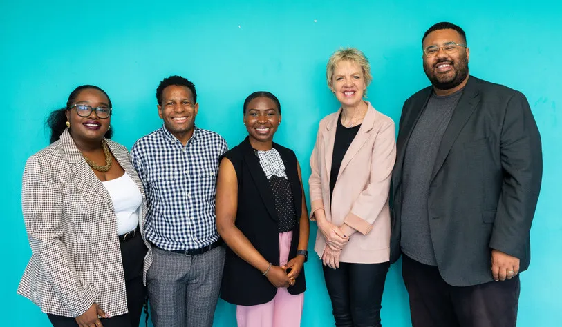 Five people stand smiling against a teal background, exuding a warm, welcoming atmosphere. They are dressed in business casual attire.
