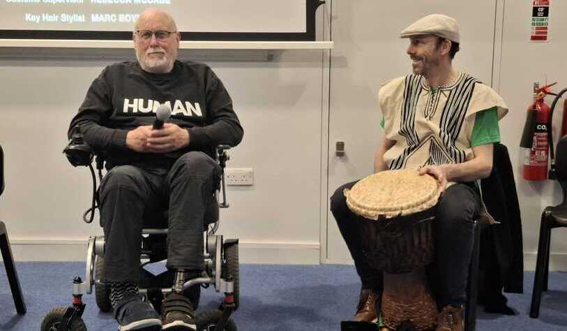 A man in a wheelchair holds a microphone, wearing a 