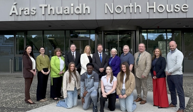 A group of fifteen people stands in front of a building entrance with the sign “Áras Thuaidh North House” above them, posing for a group photo.