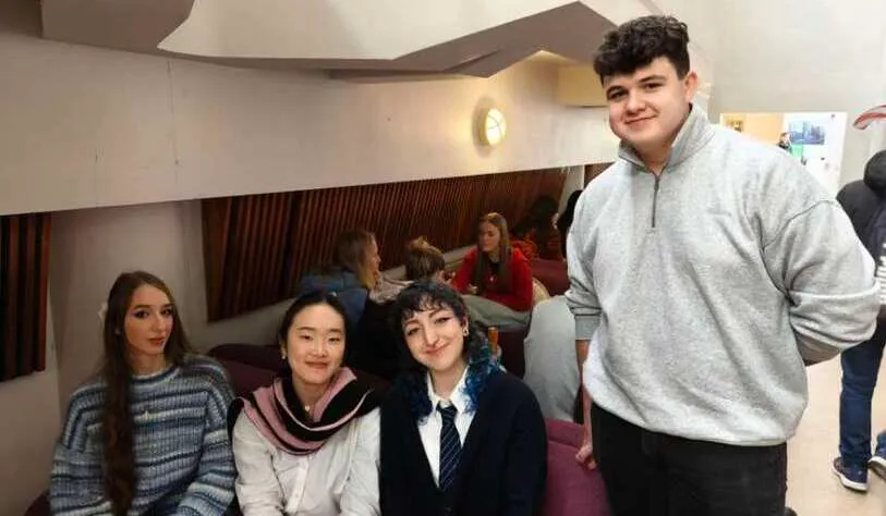 A group of four young adults sitting together, smiling in a cozy indoor setting. The atmosphere is relaxed and friendly, with casual attire.
